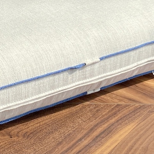 May include: A rectangular off-white cushion featuring a textured fabric surface and a prominent blue piping trim along the edge. The cushion includes a concealed zipper closure integrated into the side seam, finished with matching blue fabric detailing.