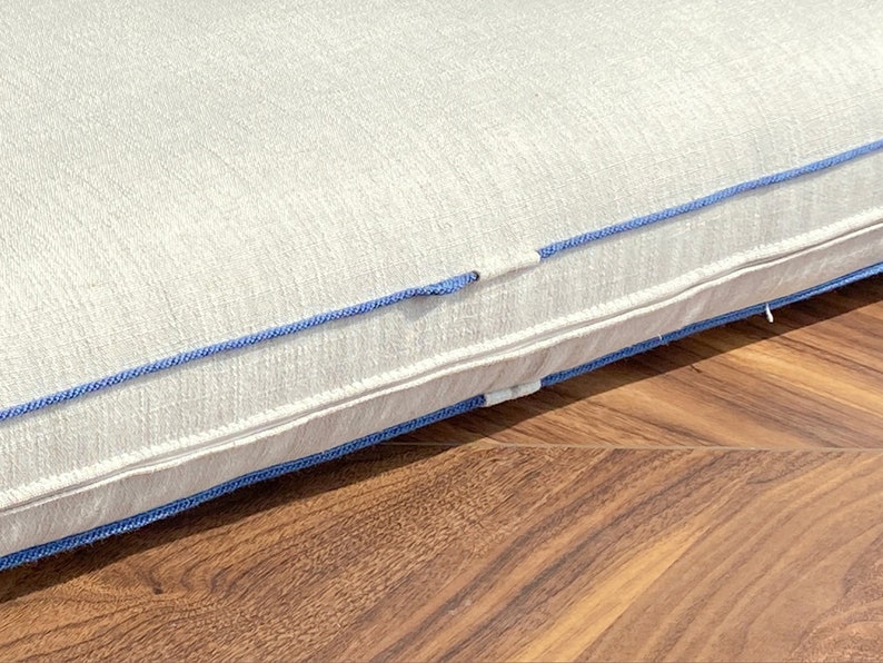 May include: A rectangular off-white cushion featuring a textured fabric surface and a prominent blue piping trim along the edge. The cushion includes a concealed zipper closure integrated into the side seam, finished with matching blue fabric detailing.