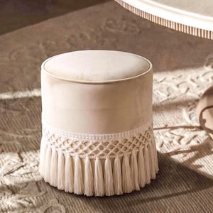 Op de afbeelding: Een cilindervormige poef bekleed met een gladde, lichtbeige stof. De onderkant van de poef is versierd met een brede band van crèmekleurig macramé vlechtwerk dat overgaat in lange, dichte franjes die tot op de grond reiken.