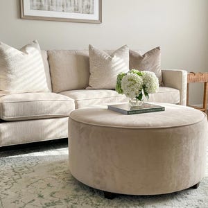 May include: A beige three-cushion sofa with three decorative throw pillows, a large round beige ottoman, and a glass vase containing white hydrangea flowers placed on a book titled Royal Gardens of the World.