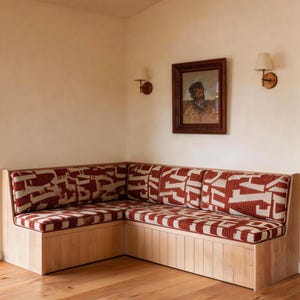 Op de afbeelding: Een ingebouwde hoekbank met een licht houten frame en rood-crèmekleurige kussens. De bank staat in een kamer met lichtgekleurde muren, hardhouten vloeren en een ingelijst portret. Twee wandkandelaars zijn aan de muur bevestigd.