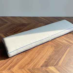 May include: A long, rectangular bench cushion featuring a light beige, textured fabric cover. The cushion is finished with a distinct, thin navy blue piping along the edges, providing a clean, structured outline to the rectangular form.