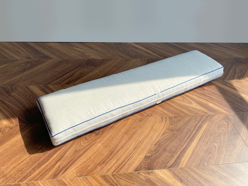 May include: A long, rectangular bench cushion featuring a light beige, textured fabric cover. The cushion is finished with a distinct, thin navy blue piping along the edges, providing a clean, structured outline to the rectangular form.