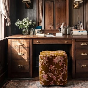 May include: A wooden desk with six drawers featuring brass pulls, a central mirror, and two wall-mounted lamps. A velvet ottoman with a brown and pink floral pattern sits in front of the desk, which holds a glass vase of white flowers and books.
