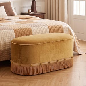 May include: An oval-shaped ottoman upholstered in mustard yellow velvet fabric. The base features a decorative trim with small pom-poms and a long, dense fringe in a matching golden-brown tone that extends down to the floor.