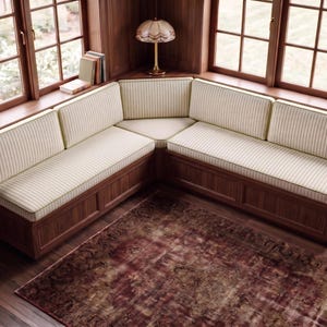 May include: An L-shaped corner bench featuring a dark wood base and cream-colored cushions with thin vertical stripes. The seating arrangement is positioned against a corner with windows, providing a structured and functional furniture piece.
