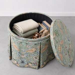 May include: A round, pleated storage ottoman upholstered in a light green fabric featuring a repeating pattern of birds, flowers, and berries. The lid is removed, revealing a black interior containing two folded blankets, two books, and a toy.