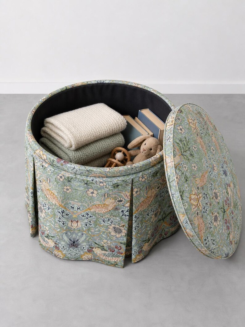 May include: A round, pleated storage ottoman upholstered in a light green fabric featuring a repeating pattern of birds, flowers, and berries. The lid is removed, revealing a black interior containing two folded blankets, two books, and a toy.