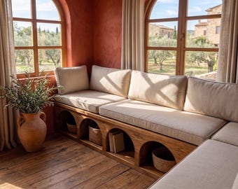 May include: A wooden bench with arched storage compartments underneath, topped with thick beige cushions. A large terracotta vase containing an olive branch sits beside the bench, which is positioned in a corner with arched windows.