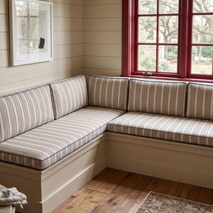 May include: An L-shaped corner bench featuring upholstered cushions with a vertical striped pattern in shades of beige, cream, and dark blue. The bench has a solid base with a smooth finish and is positioned against a wall with horizontal paneling.