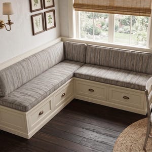 May include: An L-shaped corner bench with a cream-colored wooden base featuring built-in storage drawers with dark metal pulls. The bench includes gray and white textured fabric cushions on the seat and backrest, positioned against a wall.