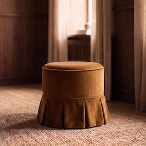Op de afbeelding: Een ronde, amberkleurige fluwelen poef met een glad, plat bovenoppervlak en een basis versierd met een geplooide stoffen rok. De kruk heeft een consistente, zacht getextureerde afwerking over de gehele cilindrische structuur voor een zitvlak.