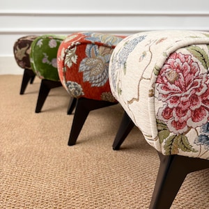 Può includere: Una fila di quattro poggiapiedi imbottiti con gambe in legno inclinate color marrone scuro. Ogni sgabello presenta un motivo in tessuto distinto, tra cui una base color crema con grandi stampe floreali rosse e blu, un motivo floreale arancione bruciato, verde e marrone.