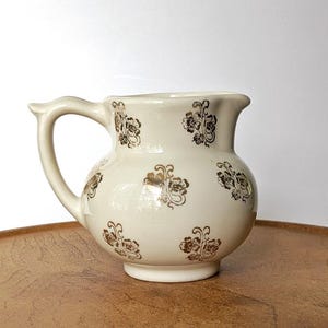 May include: A cream-colored ceramic creamer pitcher featuring a repeating pattern of small, metallic gold floral motifs. The pitcher has a rounded body, a short spout, and a curved handle extending from the side, with a smooth, glossy finish.