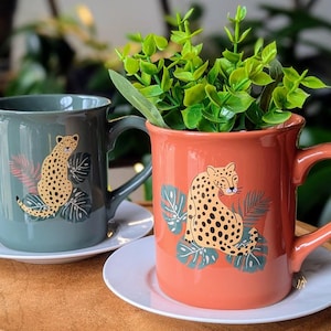 Puede incluir: Dos tazas de cerámica con temática de la jungla. Una taza es verde con un diseño de estampado de leopardo y la otra es naranja con un diseño de estampado de leopardo. Ambas tazas tienen un platillo blanco.