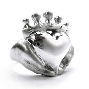 May include: A silver ring with a heart design, the heart is held by two hands, and the ring is topped with a crown.