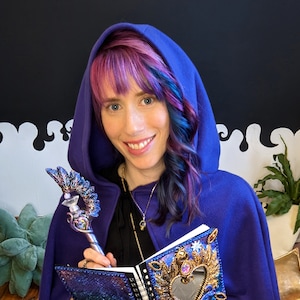 Pu&ograve; includere: Una donna con un mantello con cappuccio viola tiene un diario viola e argento con una copertina decorata. Il diario ha uno specchio a forma di cuore e decorazioni dorate. Tiene una bacchetta d'argento e viola con una punta di cristallo.