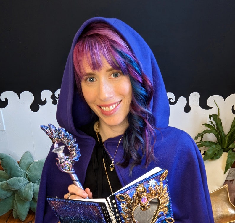 Pu&ograve; includere: Una donna con un mantello con cappuccio viola tiene un diario viola e argento con una copertina decorata. Il diario ha uno specchio a forma di cuore e decorazioni dorate. Tiene una bacchetta d'argento e viola con una punta di cristallo.