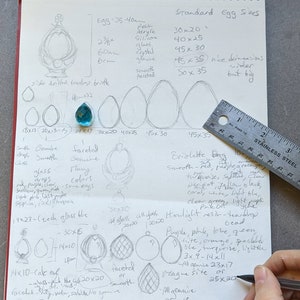 May include: Handwritten notes on a notebook page about egg sizes and types of glass beads. The notes include measurements in inches and centimeters, colors, and descriptions of the beads. The page also includes a ruler and a drawing of a teardrop-shaped briolette bead.