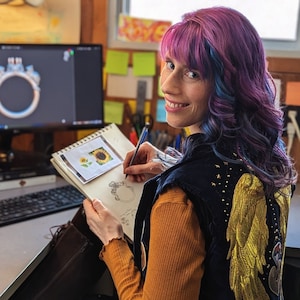 Op de afbeelding: Een vrouw met paars en blauw haar schetst een ontwerp voor een ring op een vel papier. Ze draagt een zwart vest met gouden vleugels en een geel shirt.
