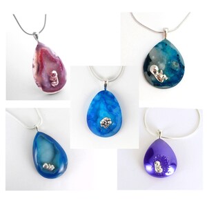 May include: Five silver necklaces with teardrop-shaped pendants. Each pendant is made of agate and features a small silver figurine of a baby inside. The pendants are in shades of blue, purple, and pink.