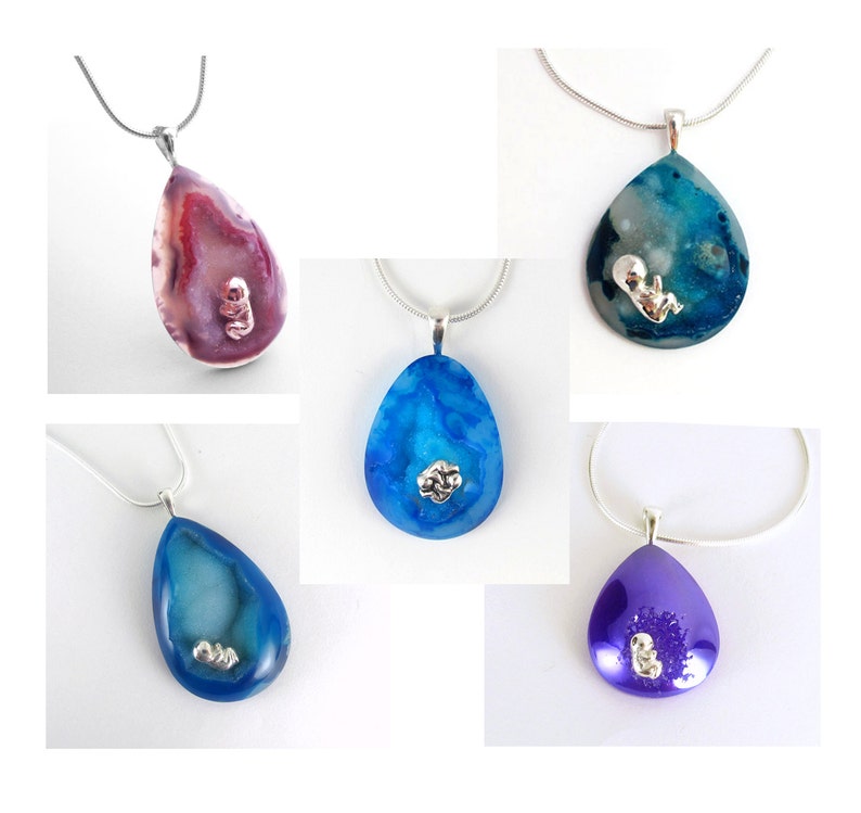 May include: Five silver necklaces with teardrop-shaped pendants. Each pendant is made of agate and features a small silver figurine of a baby inside. The pendants are in shades of blue, purple, and pink.