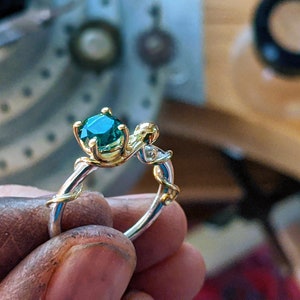Pu&ograve; includere: Un anello d'oro con una pietra preziosa verde incastonata in un design a forma di vite. L'anello &egrave; indossato al dito di una persona.