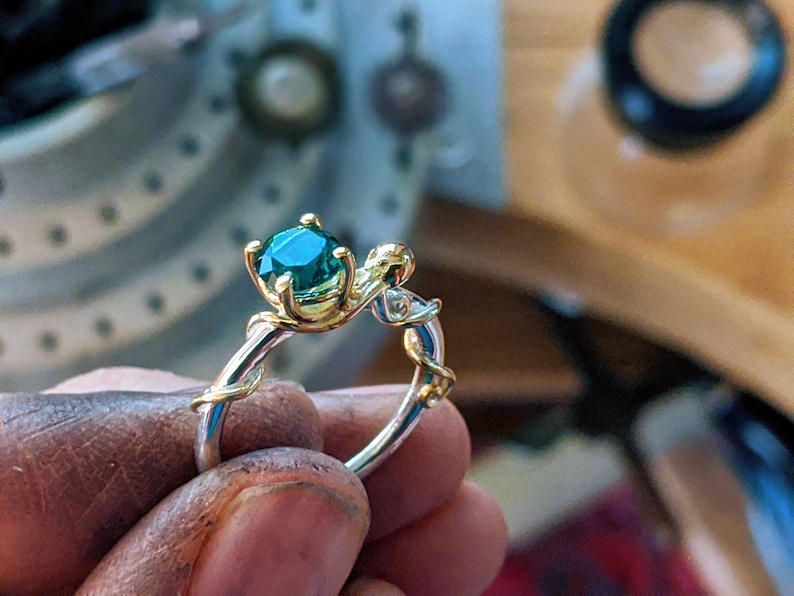 Pu&ograve; includere: Un anello d'oro con una pietra preziosa verde incastonata in un design a forma di vite. L'anello &egrave; indossato al dito di una persona.
