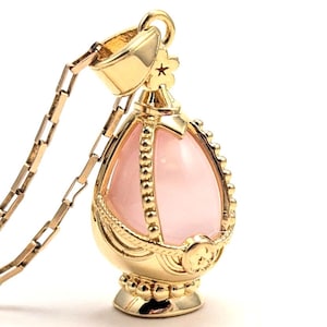 May include: A gold pendant necklace with a pink gemstone in the shape of an egg. The pendant is detailed with intricate gold designs and a small star on top.