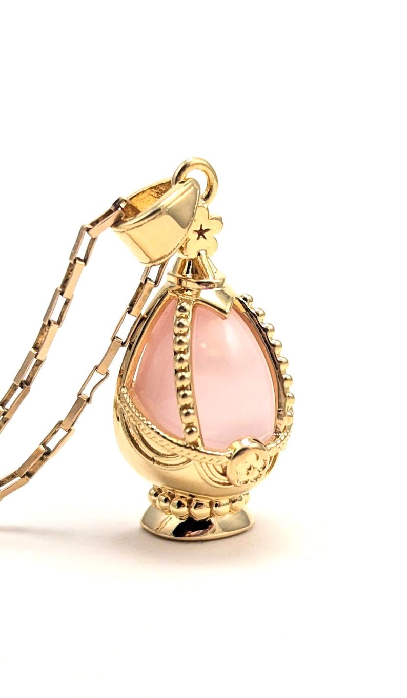 May include: A gold pendant necklace with a pink gemstone in the shape of an egg. The pendant is detailed with intricate gold designs and a small star on top.