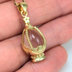 May include: A gold-tone pendant necklace features a delicate floral design and a pink quartz egg-shaped stone encased in a cage-like setting.