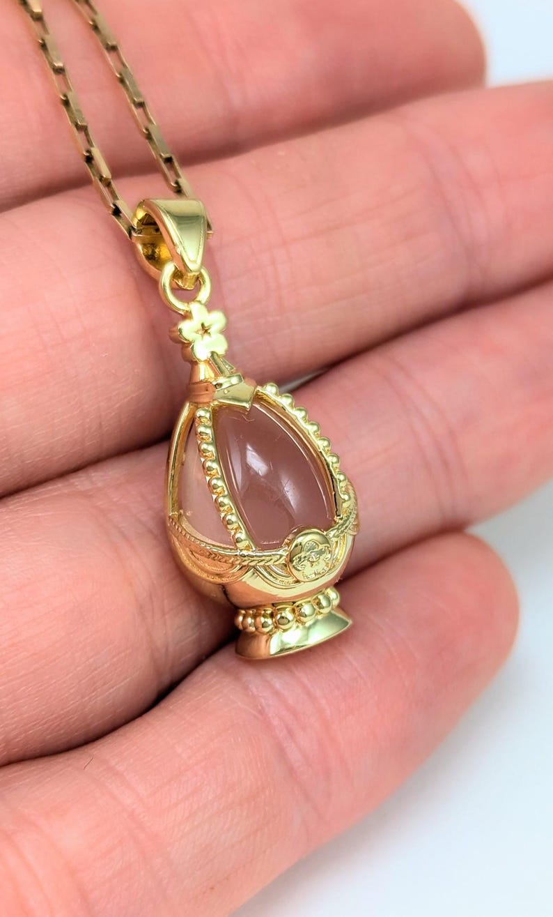 May include: A gold-tone pendant necklace features a delicate floral design and a pink quartz egg-shaped stone encased in a cage-like setting.