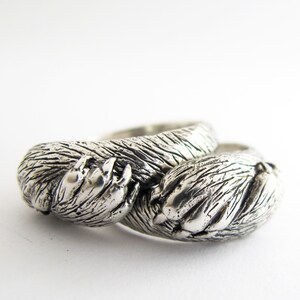 May include: Two silver rings, each shaped like a bear paw, are intertwined. The rings are detailed with textured fur and claws.