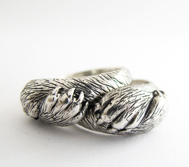 May include: Two silver rings, each shaped like a bear paw, are intertwined. The rings are detailed with textured fur and claws.