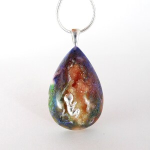 May include: A teardrop-shaped pendant necklace with a silver chain. The pendant is made of clear resin with a colourful, swirling pattern and a small silver figurine of a baby inside. The resin also contains small, white crystals.