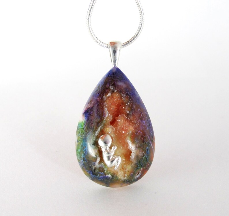May include: A teardrop-shaped pendant necklace with a silver chain. The pendant is made of clear resin with a colourful, swirling pattern and a small silver figurine of a baby inside. The resin also contains small, white crystals.