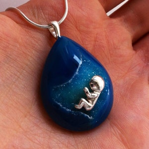 May include: A silver chain necklace with a blue agate pendant featuring a silver baby figurine inside. The pendant is shaped like a teardrop.