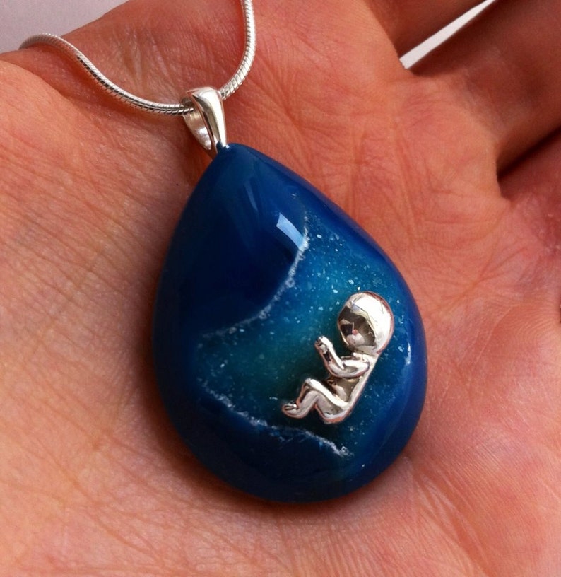 May include: A silver chain necklace with a blue agate pendant featuring a silver baby figurine inside. The pendant is shaped like a teardrop.