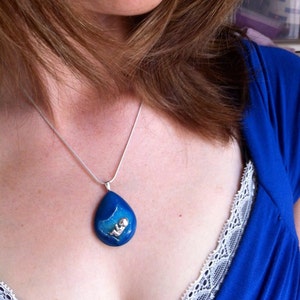 May include: A silver chain necklace with a blue teardrop pendant featuring a small silver figure inside. The pendant is made of resin and has a sparkly finish.