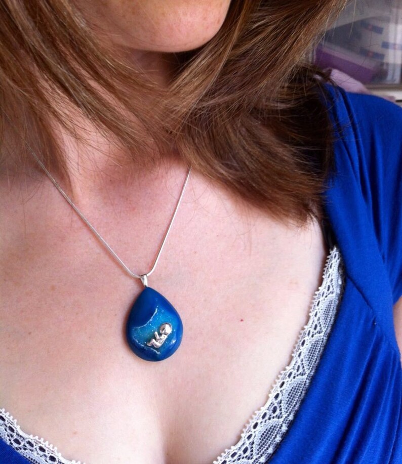 May include: A silver chain necklace with a blue teardrop pendant featuring a small silver figure inside. The pendant is made of resin and has a sparkly finish.