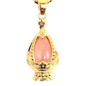 May include: A gold chain necklace with a pink gemstone pendant in the shape of an egg. The pendant has a detailed gold design.