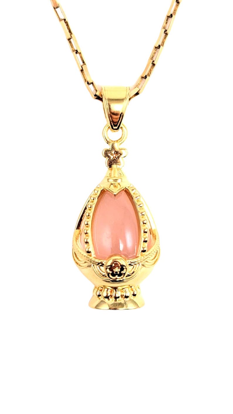 May include: A gold chain necklace with a pink gemstone pendant in the shape of an egg. The pendant has a detailed gold design.