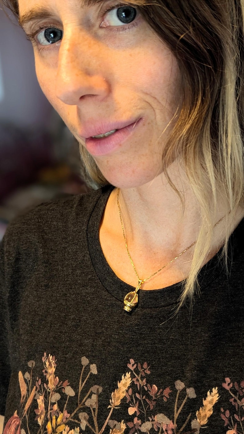 May include: A woman wearing a dark grey t-shirt with a floral print and a gold chain necklace with a small pendant.