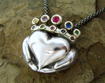 Può includere: Una collana d'argento con un ciondolo a forma di cuore tenuto da due mani. Il ciondolo è adornato da una corona di pietre preziose colorate.