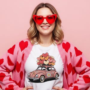May include: A woman wearing a white t-shirt with a pink car graphic carrying hearts, a pink cardigan with red heart patterns, and red heart-shaped sunglasses. The background is a solid pink color.