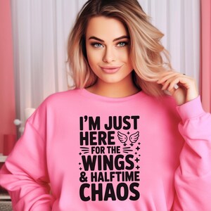 May include: A bright pink sweatshirt featuring the black text "I'M JUST HERE FOR THE WINGS & HALFTIME CHAOS". The design includes wing and star graphics. The sweatshirt has a crew neck and long sleeves.