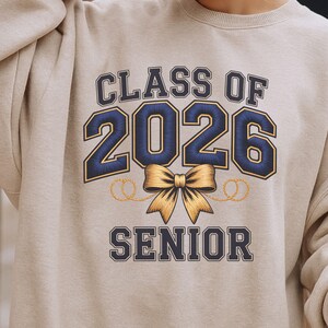 Könnte beinhalten: Beiges Sweatshirt mit dem Text "CLASS OF 2026 SENIOR" in Marineblau und Gold. Die Zahl "2026" ist das zentrale Element, mit einer goldenen Schleife und dekorativen Wirbeln darunter. Ein lässiges Kleidungsstück.