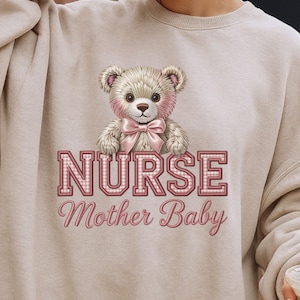 May include: A beige sweatshirt featuring a teddy bear graphic with a pink bow and accents. The word "NURSE" is printed in pink, checkered letters, with "Mother Baby" below in a script font.
