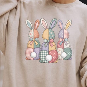 May include: A beige sweatshirt featuring a colorful graphic of three patchwork Easter bunnies. Each bunny has a different pattern, including floral, gingham, and polka dots, with a fluffy white tail and a bow tie.