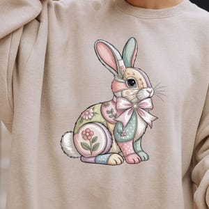 Puede incluir: Sudadera beige con un diseño de conejo de retazos colorido. El conejo tiene un gran lazo rosa, detalles florales y una variedad de colores pastel, incluyendo rosa, verde, azul y amarillo. El diseño está centrado en la parte delantera de la sudadera.